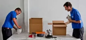 Two movers packing kitchen items into cardboard boxes indoors